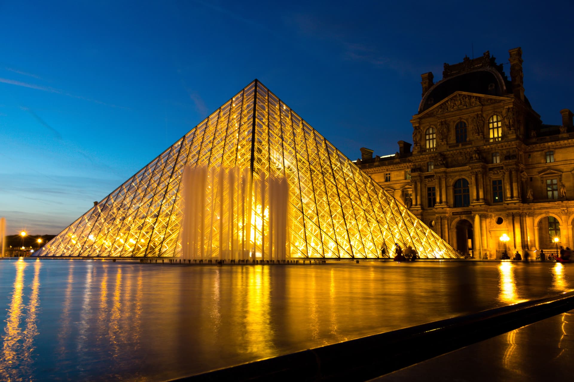 The glass pyramid of the Louvre lit gold at night, its fountain and the palace facade glowing behind