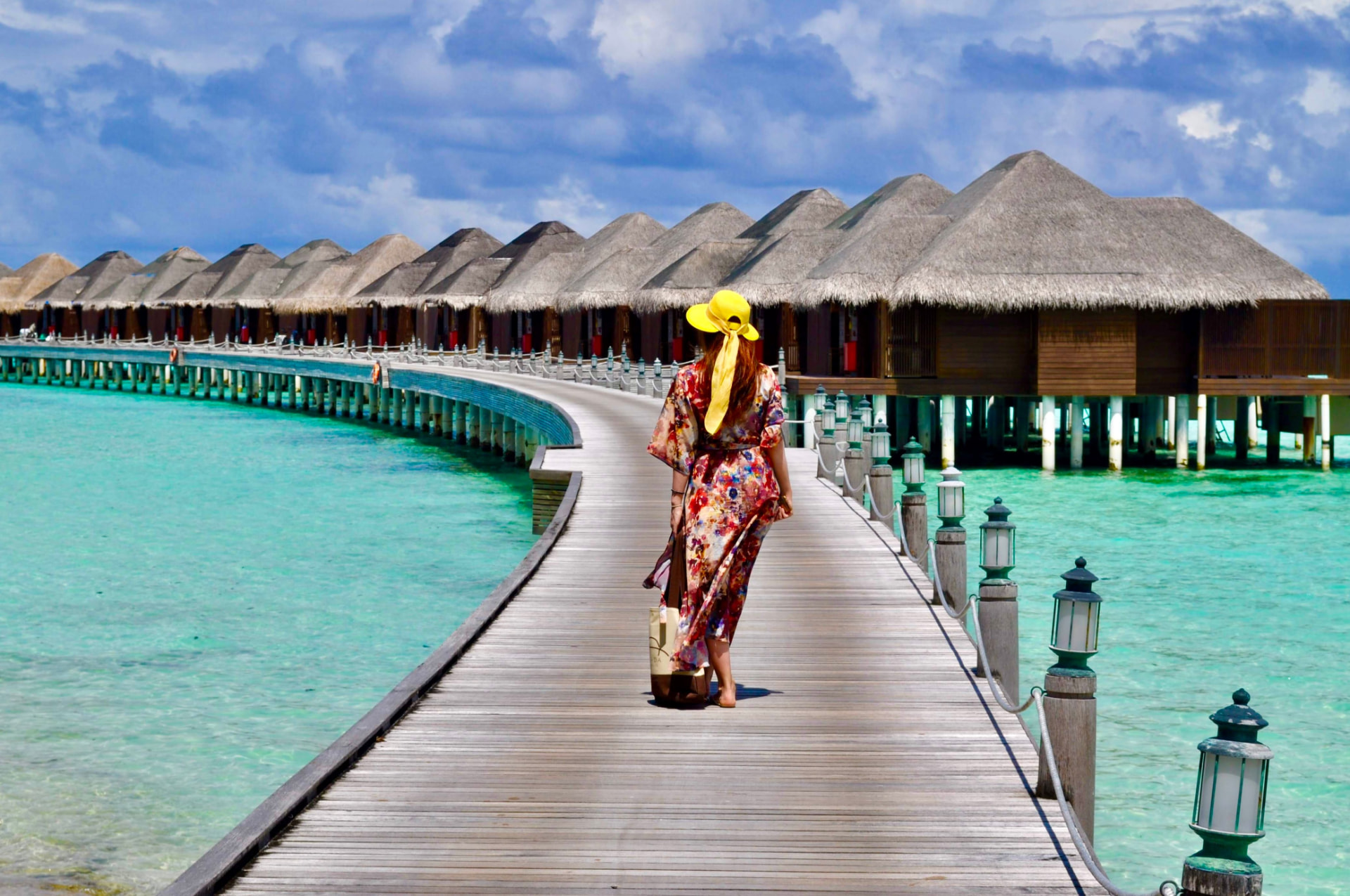 A woman in a yellow sunhat and floral dress walking along a wooden boardwalk toward a row of thatched overwater villas above a turquoise Maldives lagoon