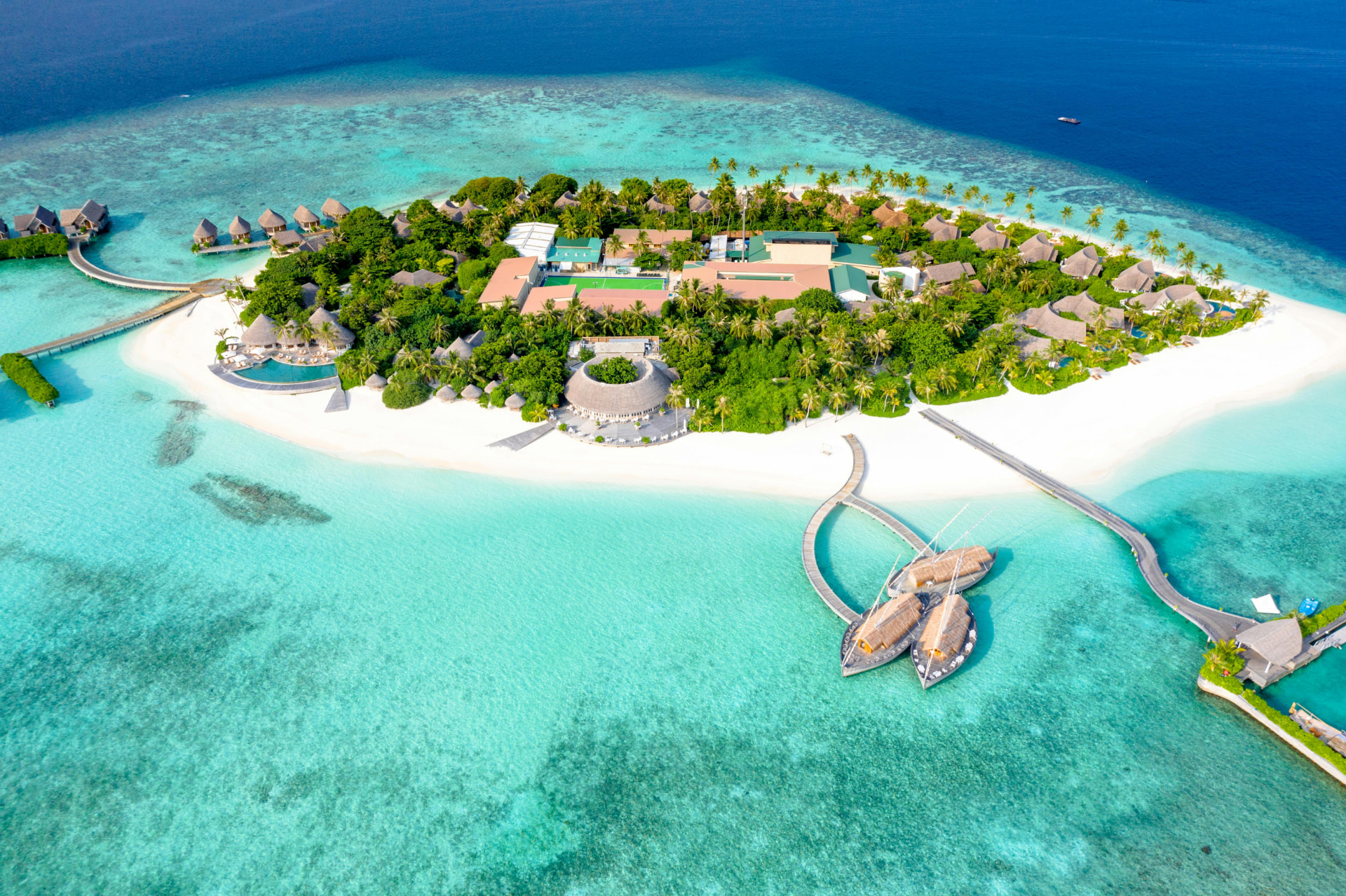 Aerial view of a private Maldives resort island ringed by white sand and a turquoise lagoon, with overwater villas on a curved boardwalk and thatched beach villas beneath palm trees
