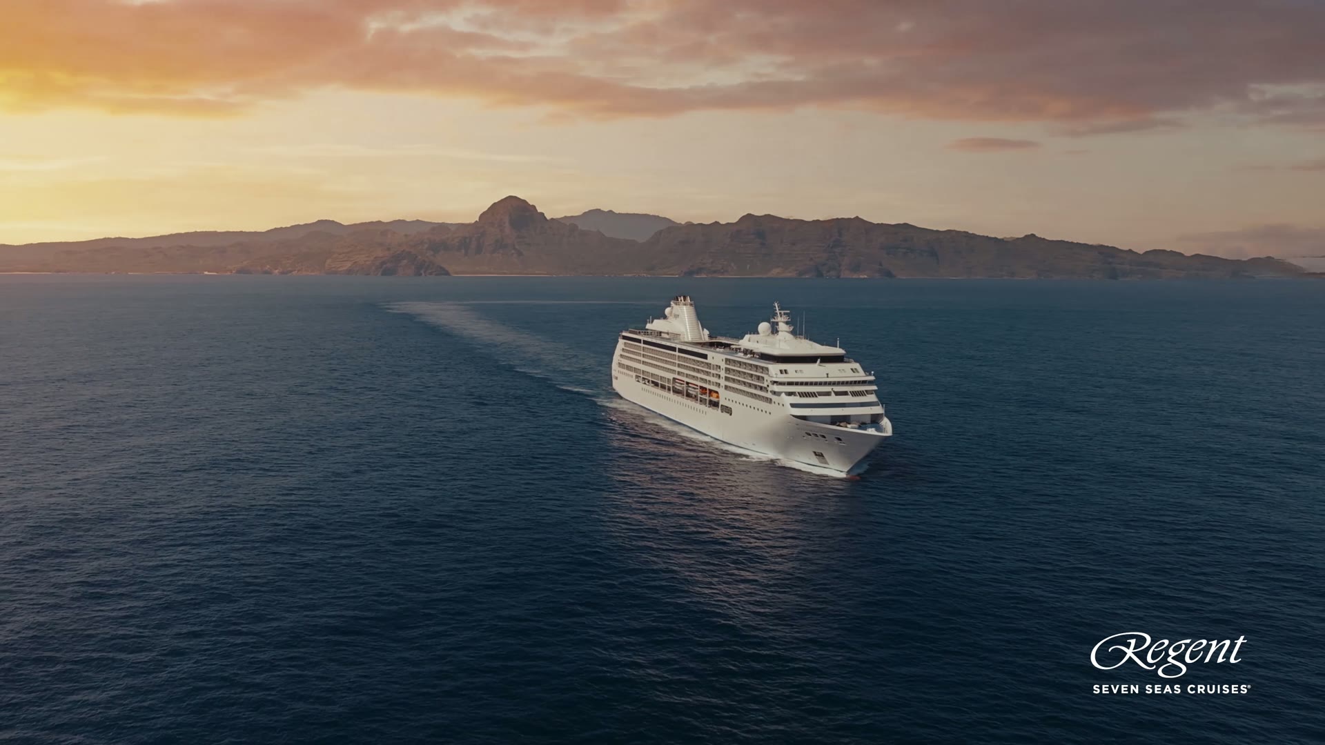A Regent Seven Seas ship sailing past a coastline at sunset
