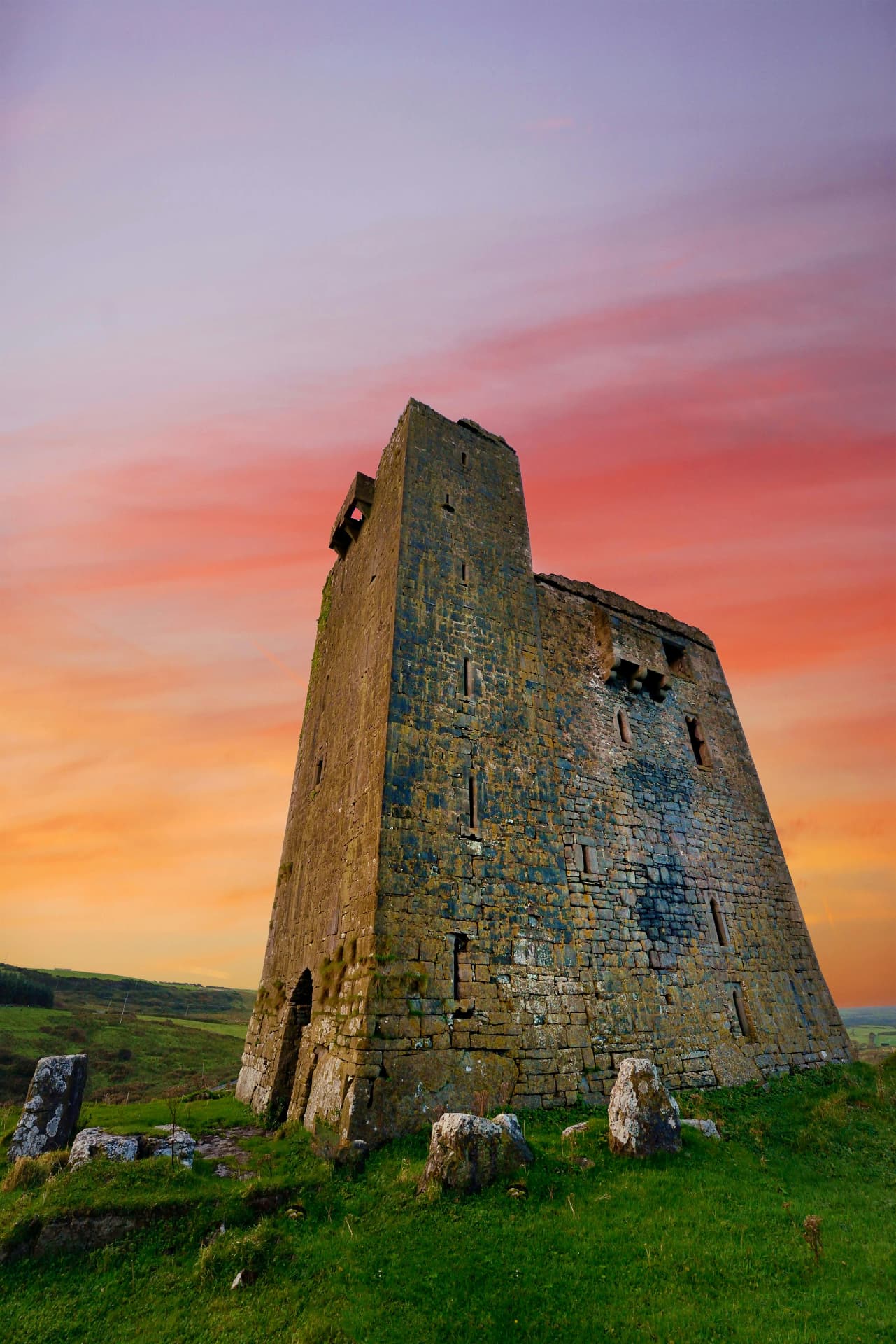 A ruined stone tower house on a grassy mound at sunset in the Irish countryside