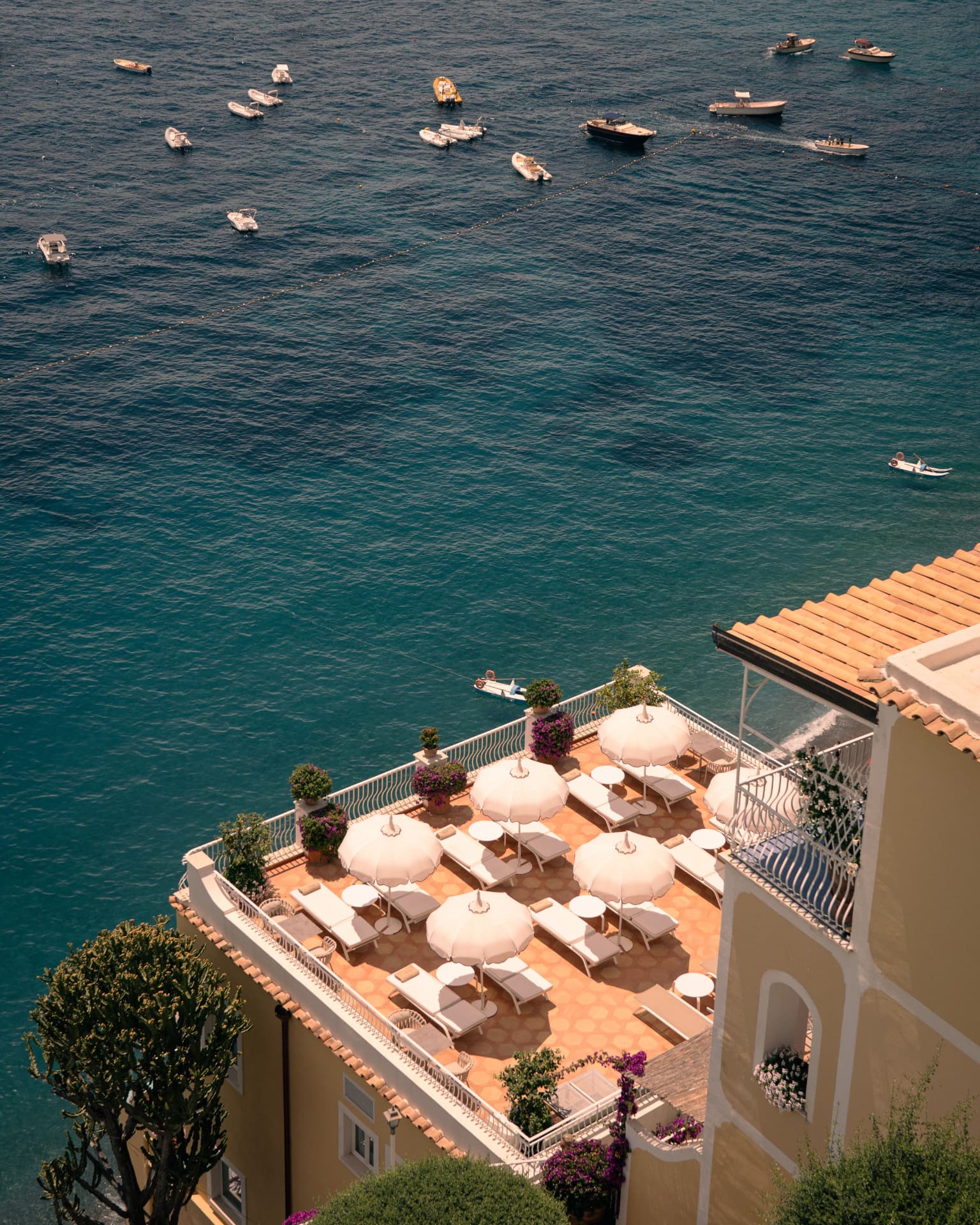 A cliffside hotel terrace above the Amalfi Coast, white parasols and loungers on warm terracotta tiles, ochre buildings and purple bougainvillea above a turquoise bay dotted with small boats